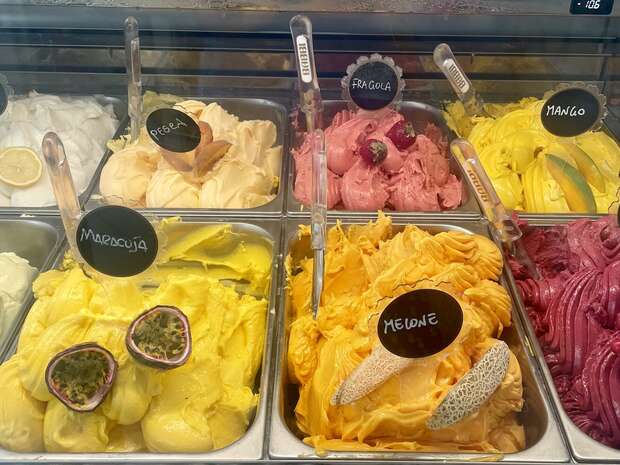 Various flavors of gelato are displayed in metal trays, topped with fruit slices. Labels read "Limone," "Pera," "Fragola," "Mango," "Maracuja," and "Melone." Scoops are inserted for serving.