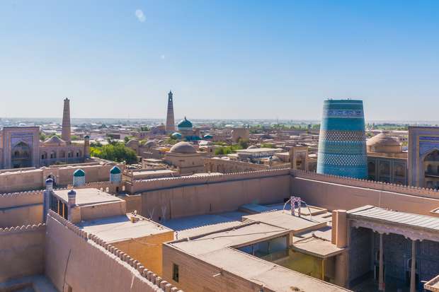 Numerous ancient buildings and minarets stand tall, showcasing intricate architectural designs, under a clear blue sky. The scene captures an expansive view of a historical cityscape with tan-colored structures.