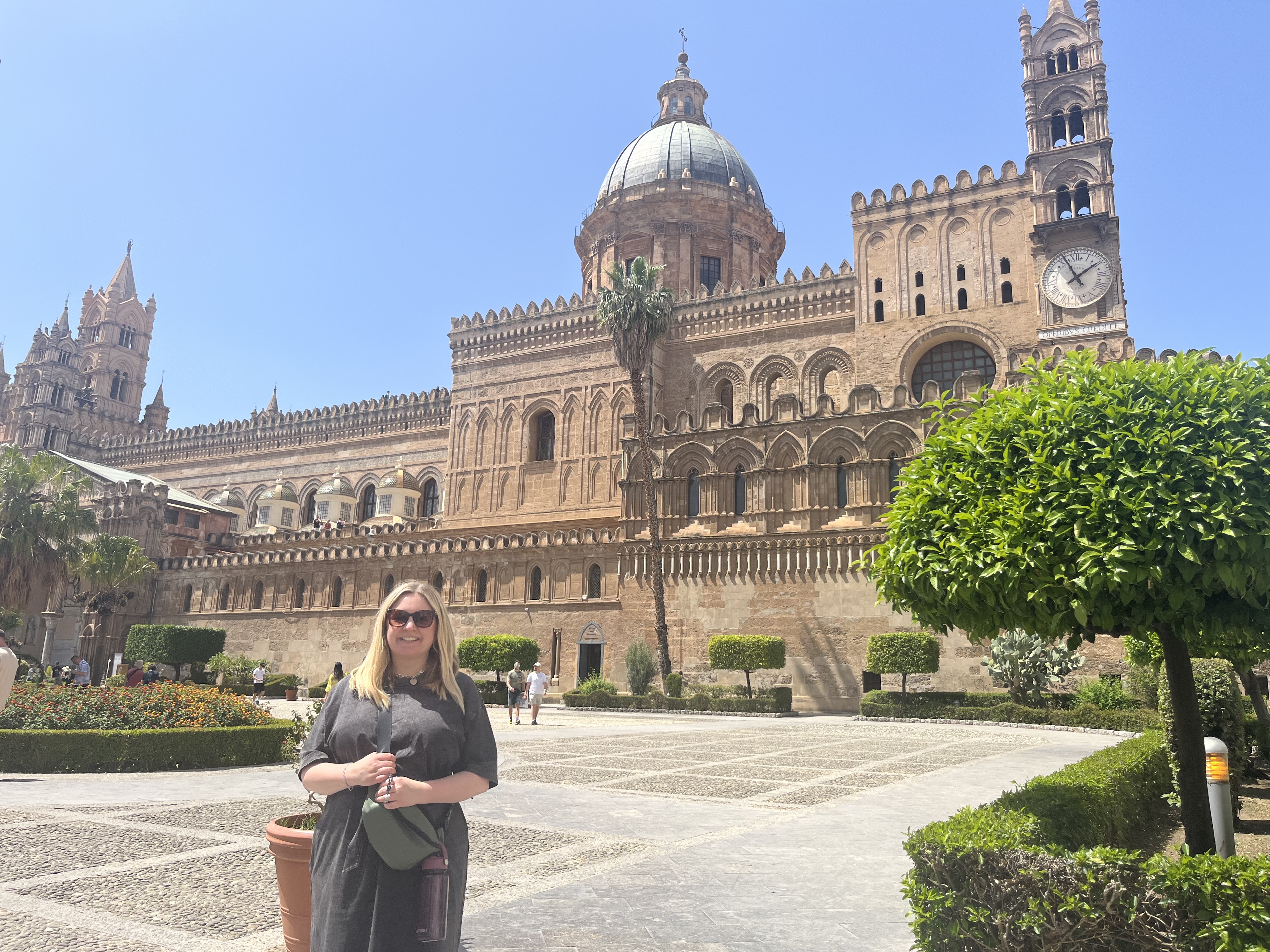 A woman stands smiling in front of a historic cathedral with intricate arches and a prominent dome. The sunny courtyard features trimmed hedges, potted plants, and a clear, blue sky overhead.