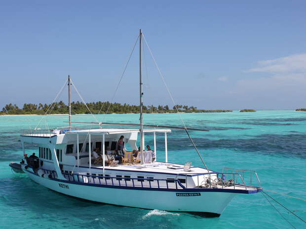 A white boat labeled "KEYFI" sails on clear turquoise water, with passengers aboard. In the background, small lush islands are visible under a clear blue sky.