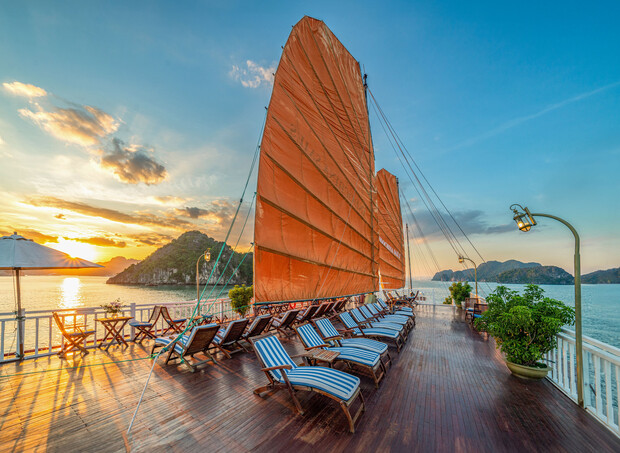 Lounge chairs with striped cushions are arranged on a wooden deck under large orange sails of a boat, set against a picturesque sunset over calm waters and distant islands.