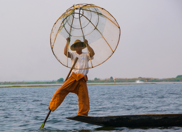 A person balances on a canoe, holding a circular fishing net above their head. They wear traditional attire, surrounded by calm waters with a distant shoreline under a clear sky.