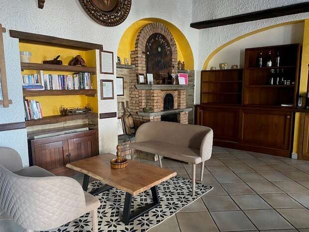 A cozy lounge features two armchairs and a wooden table on a patterned rug, with a yellow-accented bookshelf on the left and a brick fireplace in the background.
