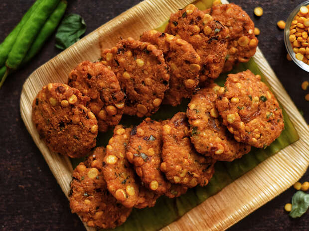 Crispy, golden-brown fritters rest neatly on a rectangular plate lined with a leaf. Nearby, small green chilies and scattered lentils add color to the dark surface.