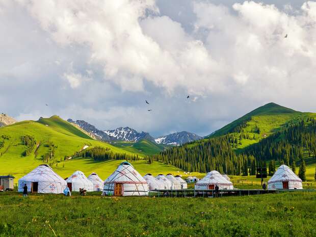 Yurts are arranged in a grassy valley with green hills and snow-capped mountains in the background. Birds are flying above under a partly cloudy sky.