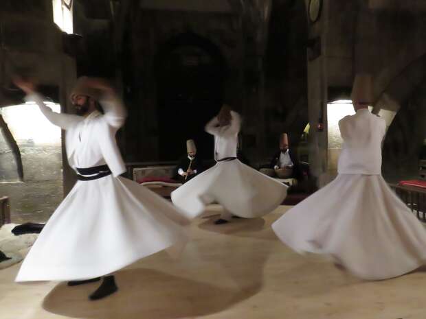 Dervishes twirl gracefully in white robes, performing a traditional dance. They are situated indoors on a wooden platform, with arched stone walls and dim lighting enhancing the spiritual atmosphere.
