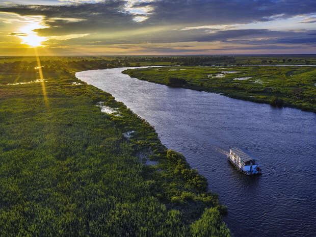 A boat travels along a winding river during sunset, flanked by lush green vegetation and marshland under a sky with scattered clouds.