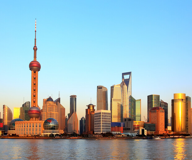 Skyscrapers, including a distinctive tower with spheres, stand illuminated by sunlight. They line the waterfront under a clear blue sky, reflecting off a calm river in an urban setting.