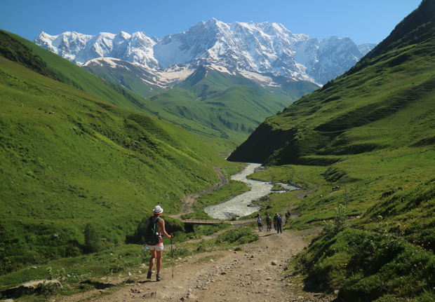 Hikers walk along a dirt path surrounded by lush green hills, with a snow-capped mountain range in the background under a clear blue sky.