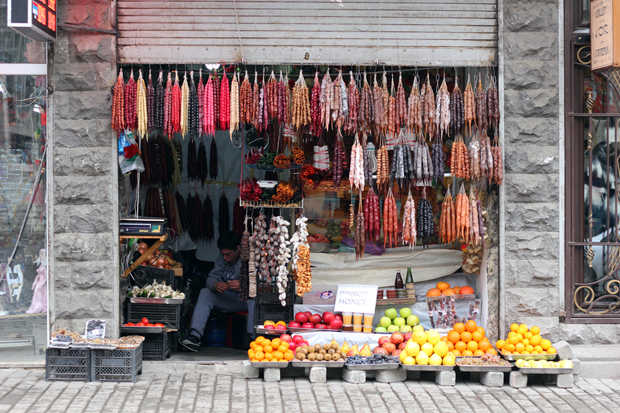 A small street shop displays hanging sausages and colorful fruits on shelves. The vendor sits inside, surrounded by goods. A sign reads, "Dr. Honey Save Save Honey."