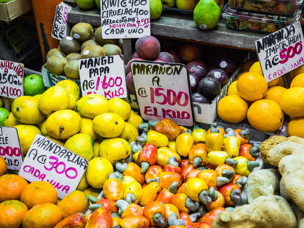 Various fruits are displayed on a market stand with handwritten prices. Signs read: "MANGO PAPA Q: 600 KILO," "MARANON A Q: 1500 EL KILO," among others. Bright, colorful arrangement.