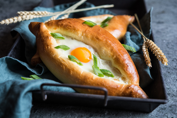 A bread boat filled with a baked egg and garnished with green onions rests on a dark cloth in a baking tray, surrounded by stalks of wheat on a rustic surface.