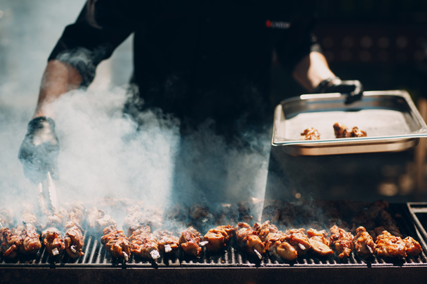 Grilled skewers are cooking on a barbecue, with smoke rising around them. A person in dark clothing and gloves holds a metal tray beside the grill in an outdoor setting.