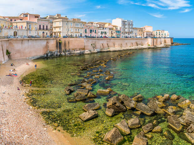 Turquoise water gently laps against a rocky shoreline beneath a long pink sea wall, lined with colorful historic buildings. People relax on the pebbled beach and enjoy the sunny weather.