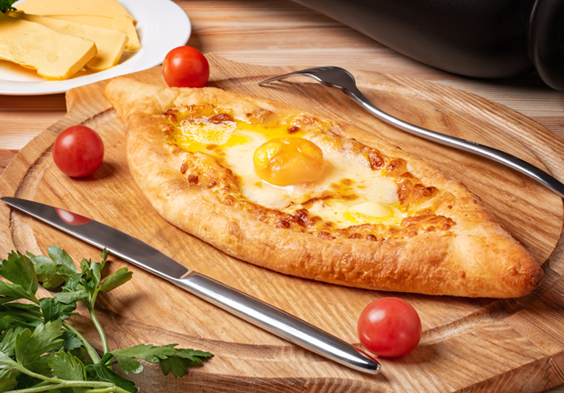 A boat-shaped bread filled with melted cheese and an egg, sitting on a wooden board, surrounded by cherry tomatoes, parsley, and cutlery. A plate of sliced cheese appears in the background.