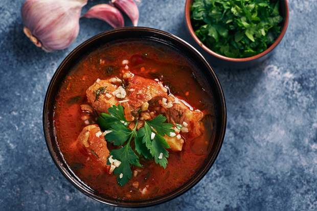 A bowl of hearty soup with chunks of meat and vegetables, garnished with fresh parsley. Nearby, there are garlic cloves and a bowl of chopped herbs on a textured surface.
