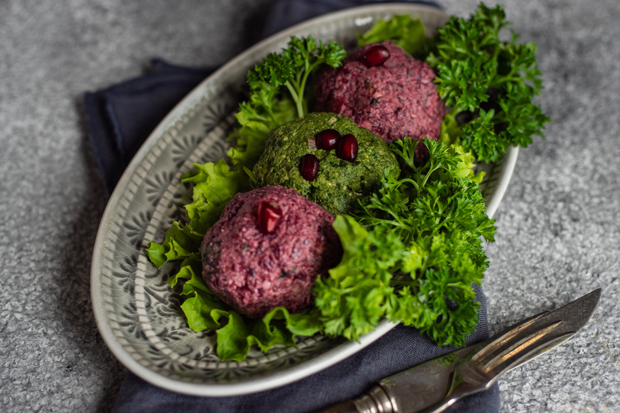 Three colorful vegetable balls garnished with pomegranate seeds sit on a bed of lettuce and parsley, arranged on an ornate oval plate atop a dark cloth near silver cutlery.
