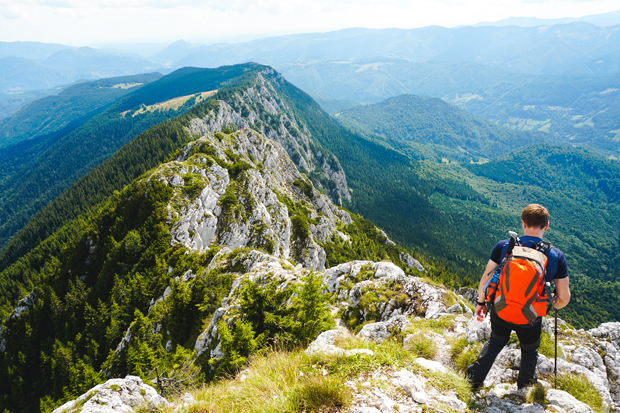 A hiker with an orange backpack navigates rocky terrain on a mountaintop, surrounded by expansive green forests and distant mountain peaks under a cloudy sky.