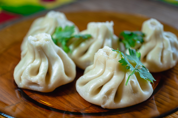 Plump dumplings rest on a wooden plate, garnished with fresh cilantro leaves. They feature pinched, pleated tops, suggesting careful craftsmanship, while bright colors blur softly in the background.