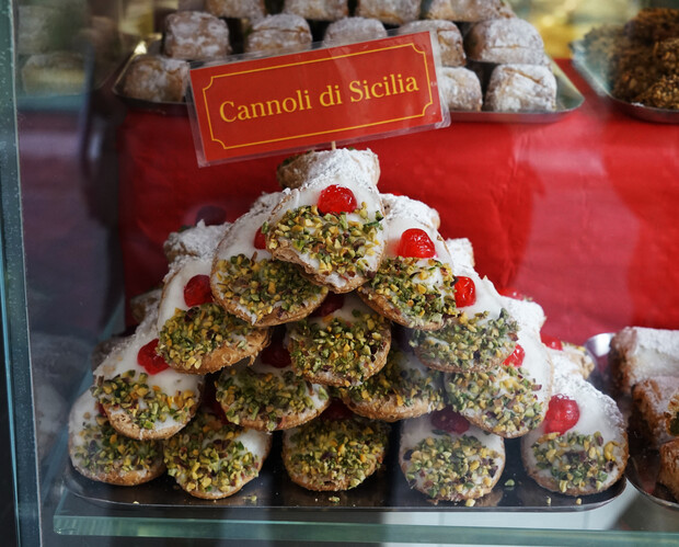 Cannoli, topped with chopped pistachios and cherries, sit stacked in a display case. A red sign above reads "Cannoli di Sicilia." Trays of sweets are visible in the background.