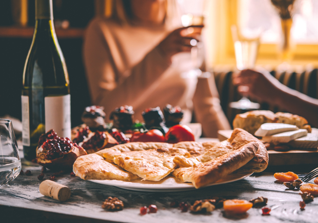 A plate of khachapuri sits on a table surrounded by wine, walnuts, dried fruits, and appetizers. Two people holding wine glasses enjoy a casual dining setting with warm, natural light.