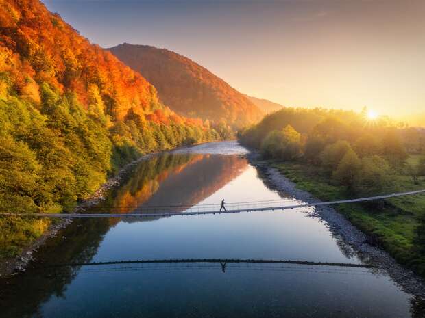 A person walks across a narrow suspension bridge over a calm river, reflecting autumn trees and a sunrise, with rolling hills in the background.