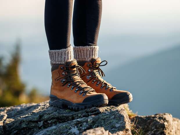 Brown hiking boots stand on a rocky surface; worn by a person with black pants and knitted socks, surrounded by a natural, blurred mountain landscape.