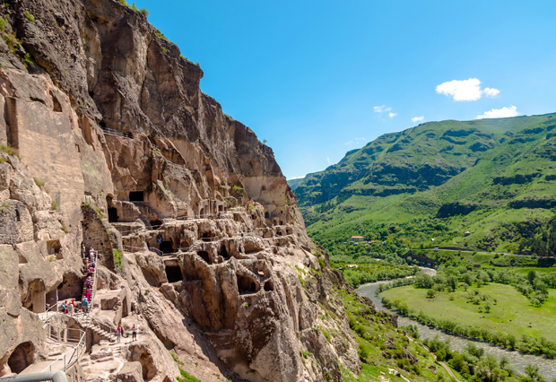 Cave dwellings carved into a rugged cliffside, with visitors exploring, overlook a lush green valley and winding river under a clear blue sky.
