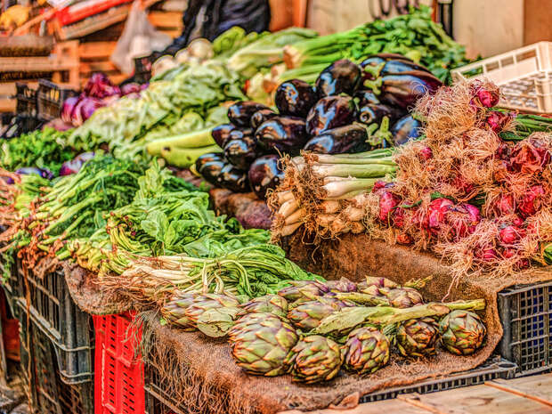 Various vegetables, including artichokes, eggplants, and onions, are displayed for sale on a market stall. Baskets and a vibrant, bustling market environment surround them.