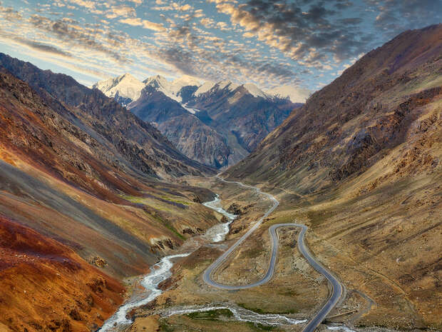 A winding road snakes through a rugged mountain valley, bordered by steep, rocky slopes. Snow-capped peaks rise in the distance under a sky streaked with clouds.