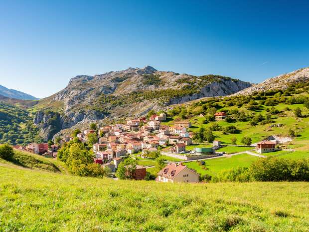 A small village with red-roofed houses sits quietly in a lush green valley, surrounded by rolling hills and rugged mountains under a clear blue sky.