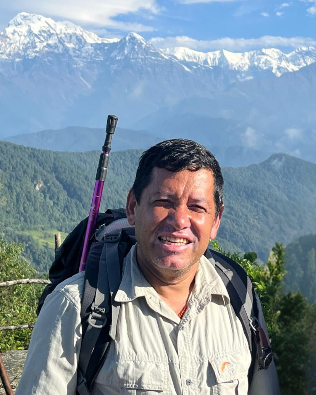 A person wearing a backpack smiles in a mountainous area, with snow-capped peaks and green hills under a clear blue sky in the background.
