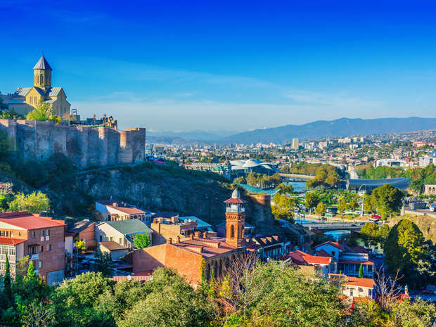 A church atop a fortress overlooks a city with vibrant buildings and trees, set against a backdrop of mountains under a clear blue sky.