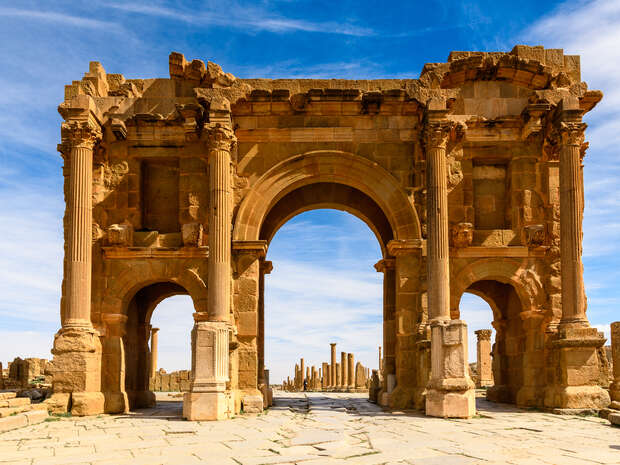 An ancient stone archway stands prominently, surrounded by scattered ruins and columns, under a bright blue sky with wispy clouds in the background.