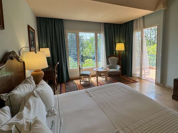 A neatly made bed with white sheets sits in a cozy room. Next to it, a table and chairs face large windows overlooking a terrace and greenery outside.