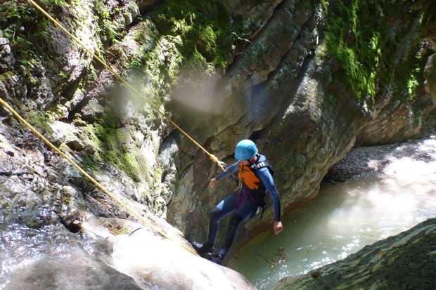A person wearing a helmet and wetsuit rappels down a moss-covered rocky canyon wall using ropes, with a pool of water at the bottom.