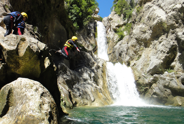 A person wearing a helmet and wetsuit leaps from a rocky ledge into turquoise water. Surrounding the scene is a canyon with a cascading waterfall and lush green foliage.