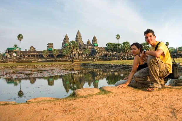 Two people sit on a dirt path by a reflective water body, with the ancient stone towers of Angkor Wat in the background, surrounded by palm trees under a clear sky.