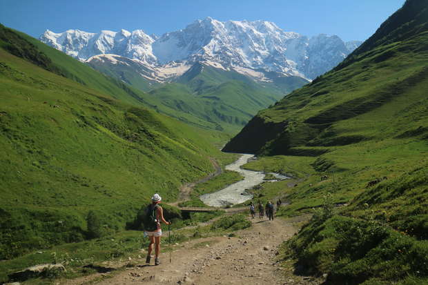 A person with a backpack walks along a dirt path in a lush, green valley, while others follow. Snow-capped mountains rise majestically in the distance under a clear blue sky.