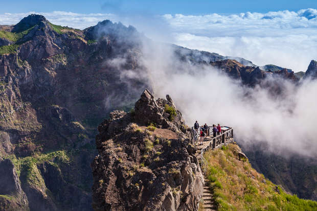 A group of people stands on a rocky cliff lookout, surrounded by clouds, with steep, green-covered mountains in the background, creating a dramatic, elevated scenic view.