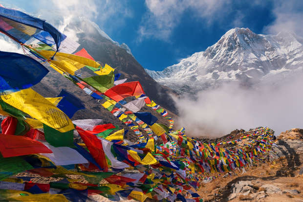 Colorful prayer flags flutter in the wind, strung along a rocky trail. Snow-capped mountains tower in the background under a bright blue sky, creating a serene alpine landscape.