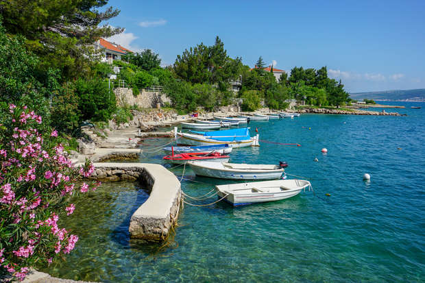 Boats float near a stone walkway beside a tranquil, clear blue sea. Pink flowers and lush greenery line the coast, while several white houses are visible through the trees.