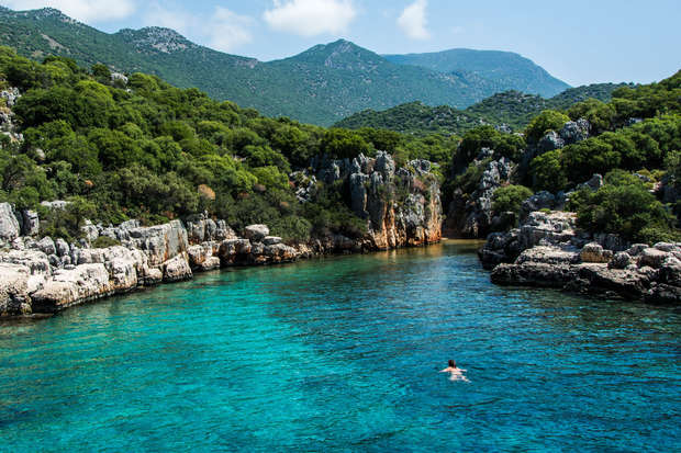 A swimmer moves through clear turquoise water in a rocky, forested lagoon surrounded by lush greenery and distant mountains under a blue sky.