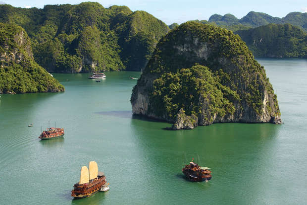 Traditional wooden boats with sails float on a calm green bay, surrounded by towering limestone karsts and lush vegetation, creating a serene, natural landscape.