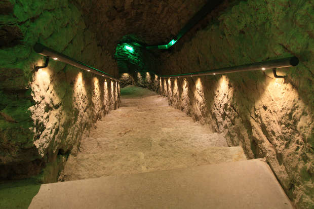 Stone staircase descends into an underground tunnel, illuminated by warm, yellow wall-mounted lights. Green ambient lighting gives the rough stone walls a mysterious and eerie atmosphere.