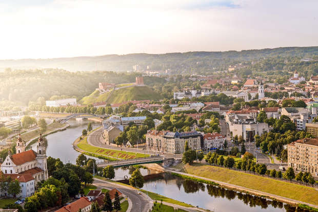 A cityscape shows a river winding through Vilnius, surrounded by historic buildings and lush greenery, with a castle on a hill in the distance under a clear sky.