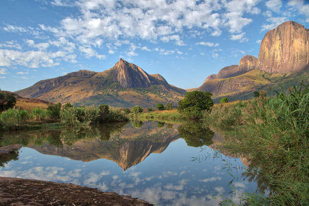 Mountains stand majestically, reflected in a calm, clear pond surrounded by lush greenery. Fluffy clouds fill the blue sky, enhancing the tranquil, picturesque landscape.