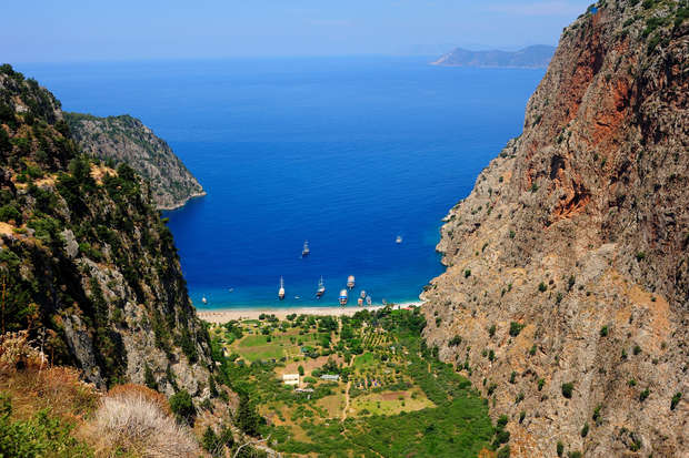 A deep blue bay framed by towering cliffs features small boats sailing. Below, a lush green valley leads to a narrow sandy beach, with hills and further mountains in the distance.
