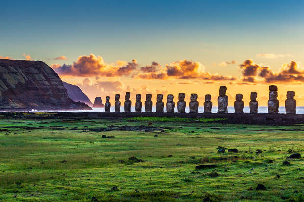Stone statues stand in a row on a grassy plain at sunset, backed by a rocky coastline and vibrant, cloud-streaked sky.