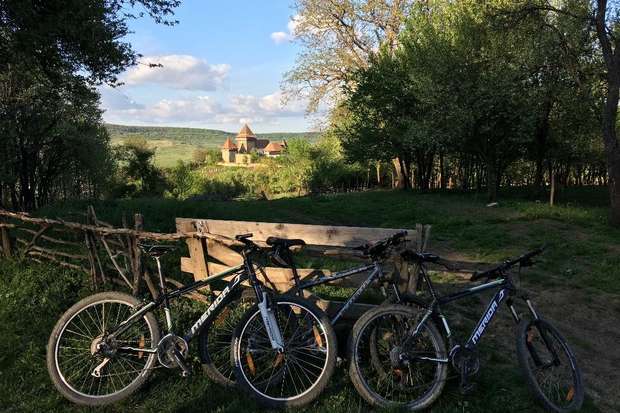 Bicycles rest against a rustic wooden fence in a lush, wooded area. In the distance, a castle-like structure stands atop a hill under a partly cloudy sky.